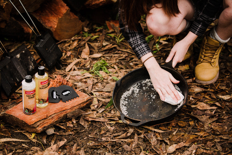 A person kneels on leafy ground, scrubbing cast iron cookware with the Campfire Cast Iron Scrub. Bottles of seasoning oil, Australian rock salt, and cleaning tools are arranged on a wooden plank nearby.