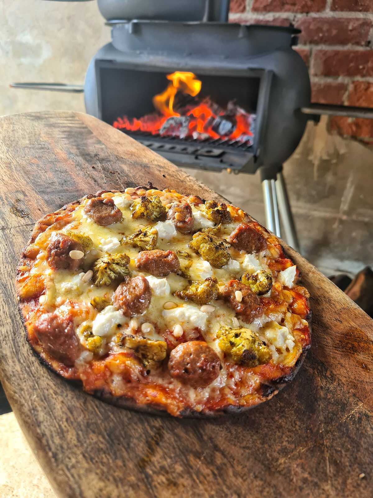 A freshly baked pizza topped with sausage and cheese rests on a wooden board in front of a wood-fired oven with visible flames, its crispy crust perfectly achieved using the Ozpig Oven Smoker 9" Pizza Stone by Ozpig.