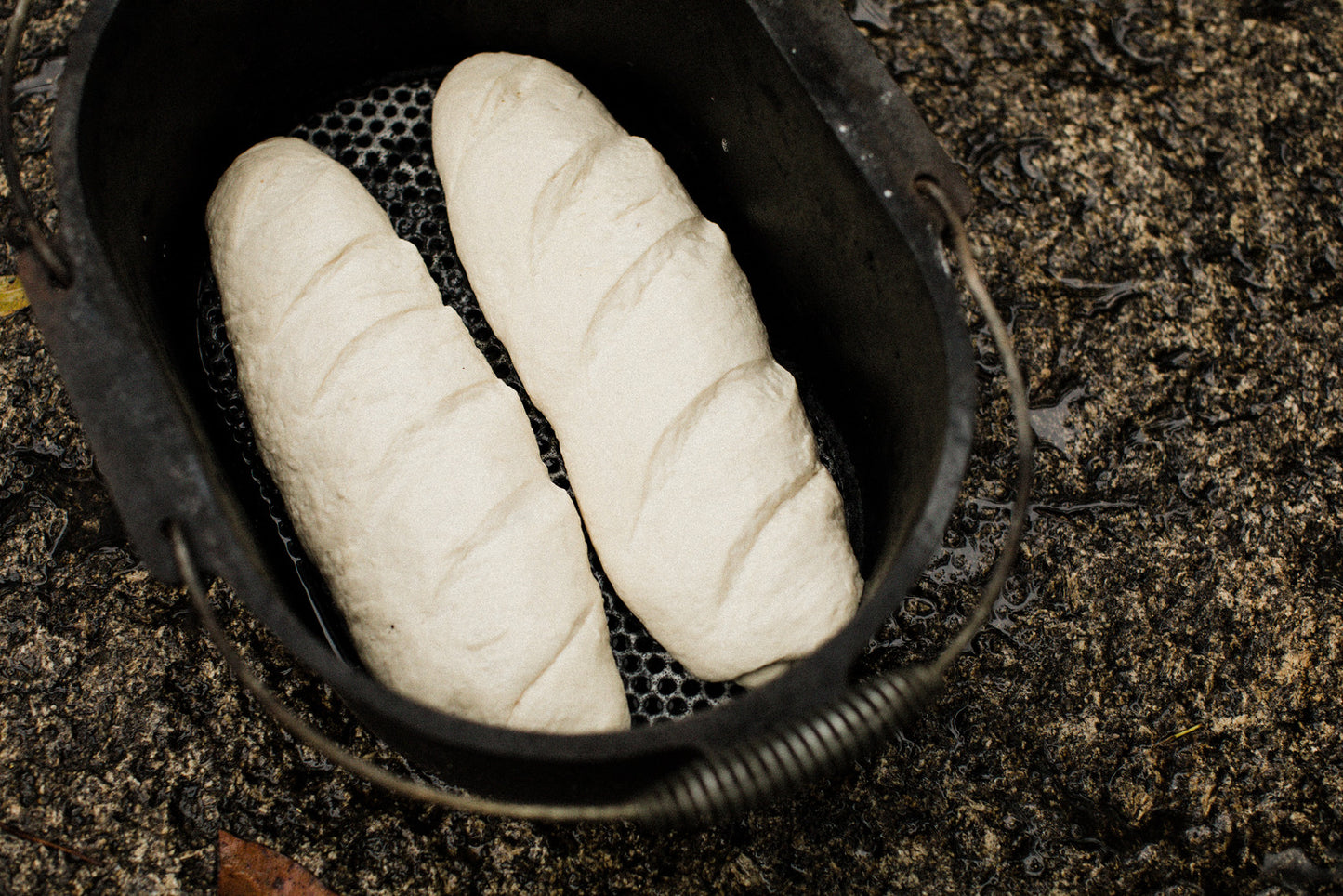 Two unbaked loaves of bread dough rest side by side in a Campfire 10 Quart Oval Camp Oven, its black cast iron construction positioned on a rough, wet outdoor surface—making it perfectly suited for campfire cooking or use as a camp oven.