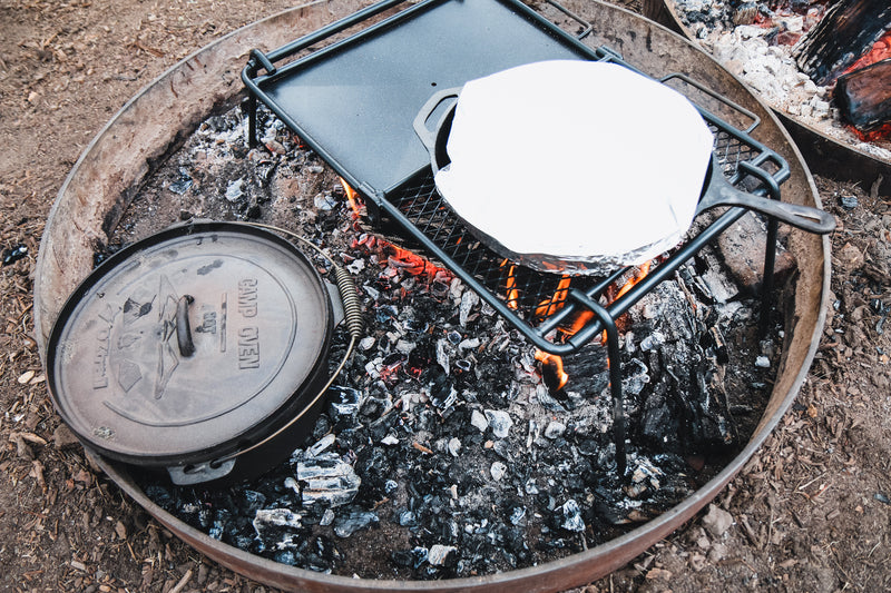Surrounded by a fire ring outdoors, hot coals heat the Campfire 4.5 Quart Camp Oven and a grill grate with a skillet covered in foil, creating the perfect setting for outdoor cooking.