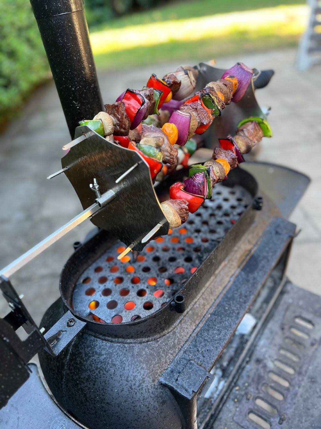 Vegetable and meat skewers are being grilled outdoors on the Ozpig Kebab Rotisserie Set by Ozpig, with stainless steel skewers over glowing coals and blurred greenery in the background.