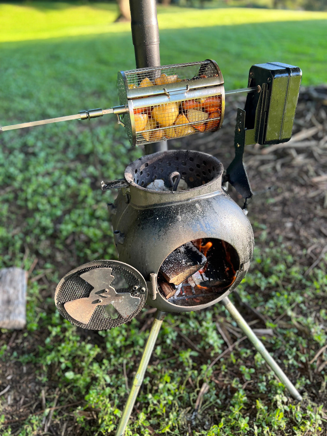 The Ozpig Rotisserie Basket Small by Ozpig cooks potatoes evenly in a rotating stainless steel basket over a wood fire, set on tripod legs with a round firebox, perfect for outdoor use in your grassy yard.
