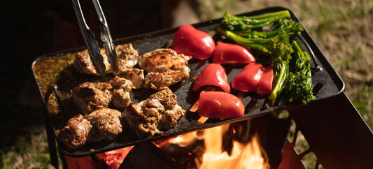 Chicken and vegetables grilling over an open fire on the Ozpig 3-in-1 Flat Pack Fire Pit, turned with tongs.