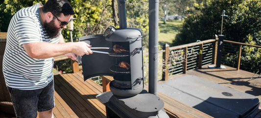 A man tends food in an Ozpig Big Pig with smoker attachment, cooking on racks in an outdoor deck setting.