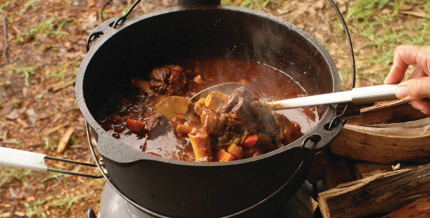 Camp oven stew simmering in a cast iron pot over a fire, being stirred with a ladle outdoors.