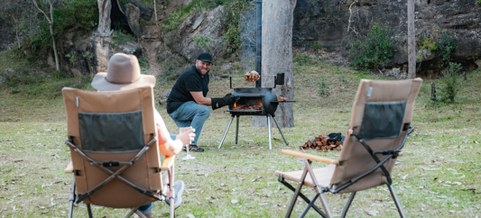 A man tends a rotisserie on an Ozpig Big Pig while two people relax in chairs in a grassy, wooded campsite.