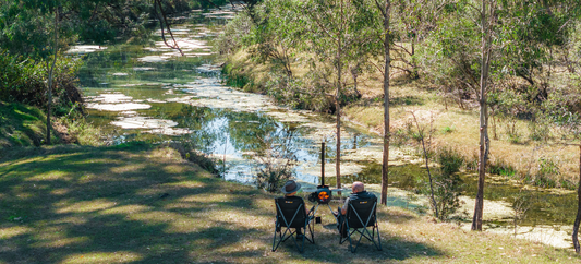 Two campers relax by a creek with an Ozpig Series 2, enjoying a peaceful, natural campsite scene.