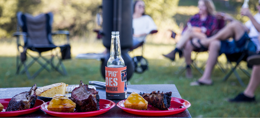 A barbecue feast sits by a smoker as people relax in lawn chairs enjoying an outdoor meal together.