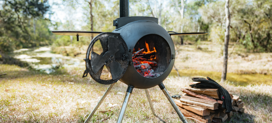 Ozpig Series 2 portable wood stove burning beside firewood and gloves, set in a grassy spot near water.