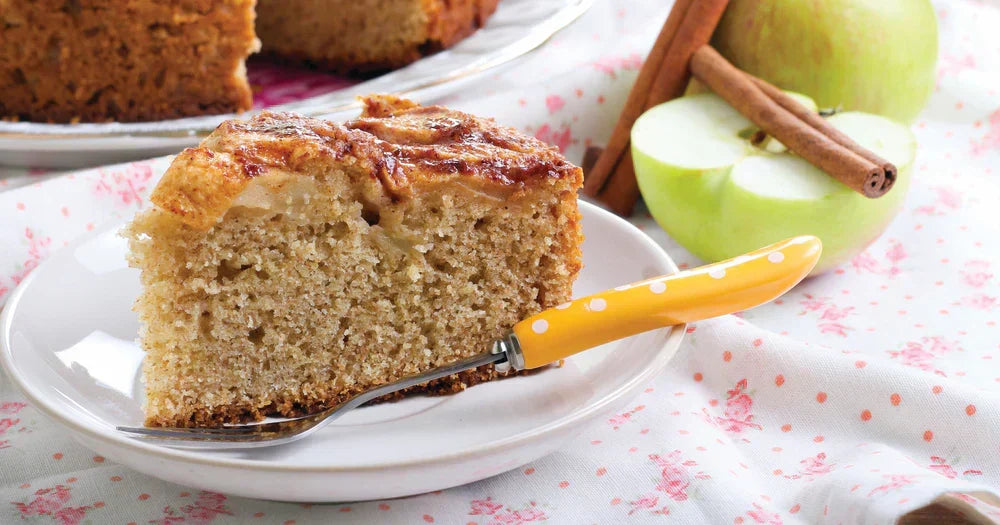 Assunta’s Apple Tea Cake slice on a plate, showing a soft crumb and baked apple topping with fresh apples and cinnamon beside it.