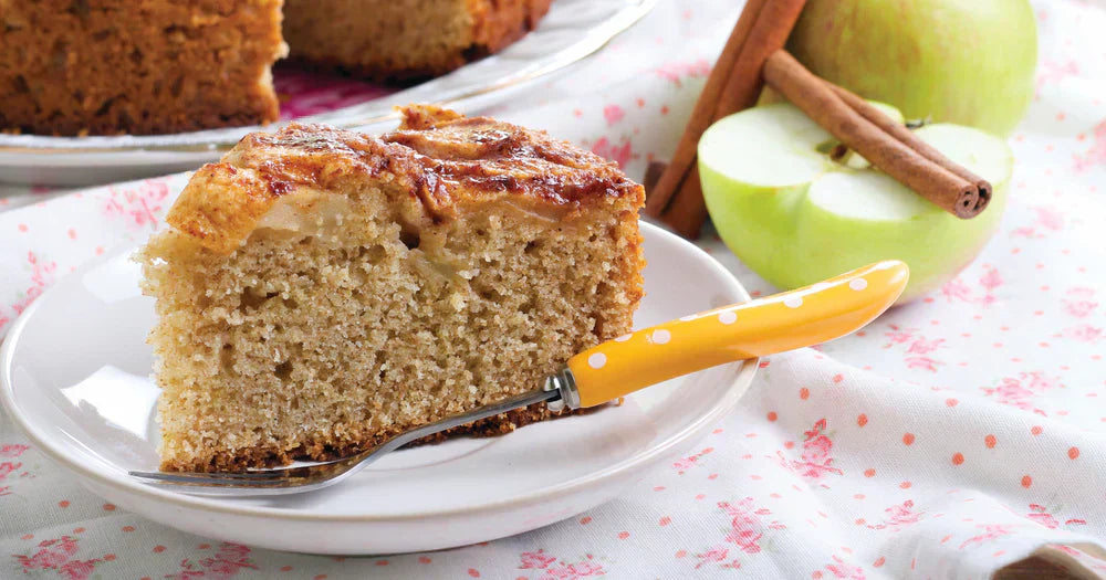 Assunta’s Apple Tea Cake slice on a plate, showing a soft crumb and baked apple topping with fresh apples and cinnamon beside it.