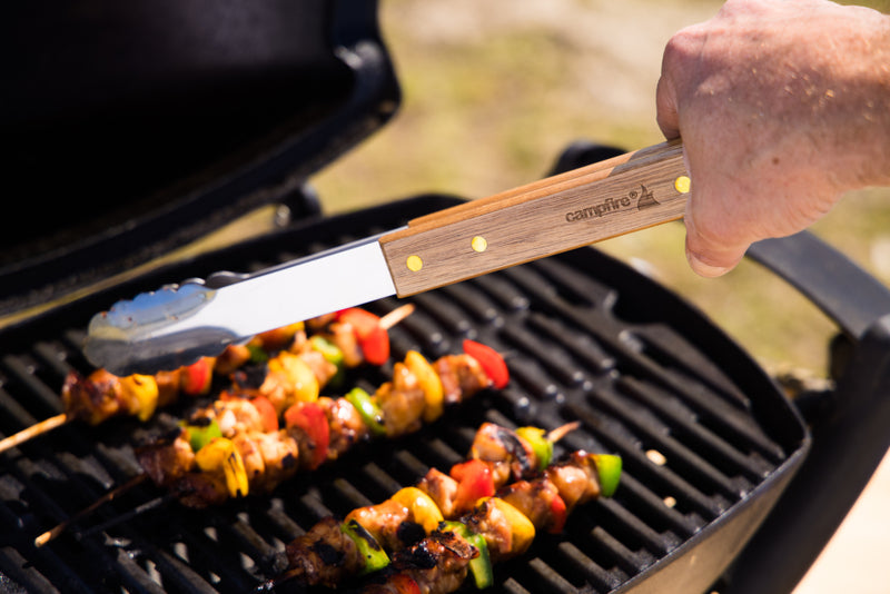 A hand uses wooden-handled tongs from the Campfire 22pc BBQ Set to grill colorful vegetable and meat skewers on a black barbecue grill during an outdoor cooking session.