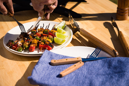 Hands serve grilled vegetable and meat kebabs on a plate with lime wedges, next to the Campfire 22pc BBQ Set of stainless steel utensils, a blue napkin, and wooden tongs on a wooden table in sunlight—perfect for outdoor cooking.