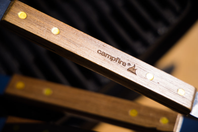 Close-up of the wooden handle of the Campfire 22pc BBQ Set, engraved with the brand name and a small flame logo, featuring brass rivets; in the background, a blurred stainless steel grill or grate—perfect for outdoor cooking—is visible.