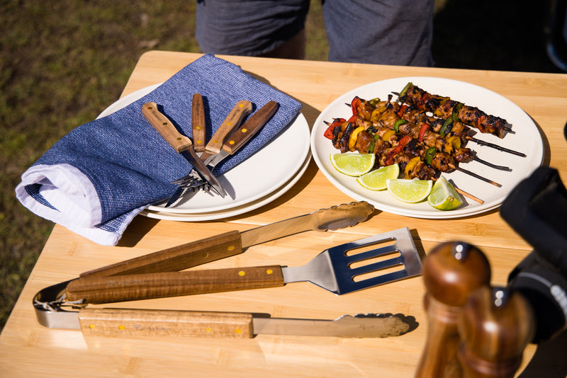 A wooden table set with grilled vegetable and meat skewers, lime wedges, blue napkins, a pepper grinder, and utensils from the Campfire 22pc BBQ Set, all ready for an outdoor cooking meal.