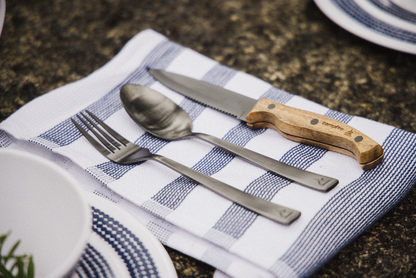 A fork, spoon, and knife from the Campfire 12pc Cutlery Set, featuring stainless steel with acacia wood handles, are arranged on a white napkin with blue stripes atop a stone surface beside a patterned plate.