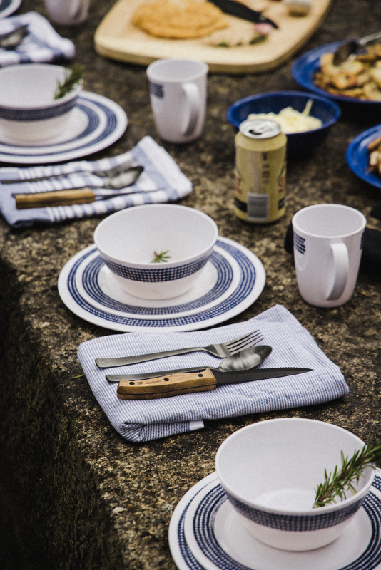 Enjoy al fresco dining with the Campfire 12pc Cutlery Set, featuring stylish acacia wood handles. Each stainless steel utensil gleams next to white bowls containing a sprig of rosemary, complemented by mugs, plates, and striped napkins. A can and an array of food dishes complete the inviting outdoor table setting.