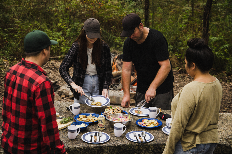 Four people, dressed casually in caps and jackets, gather around a stone picnic table outdoors with plates of food and mugs, preparing to eat with the Campfire 12pc Cutlery Set, which features stylish acacia wood handles. A campfire glows warmly in the background as they enjoy their meal using these durable stainless steel utensils.