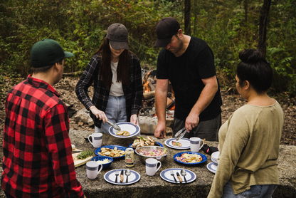 Four people, dressed casually in caps and jackets, gather around a stone picnic table outdoors with plates of food and mugs, preparing to eat with the Campfire 12pc Cutlery Set, which features stylish acacia wood handles. A campfire glows warmly in the background as they enjoy their meal using these durable stainless steel utensils.
