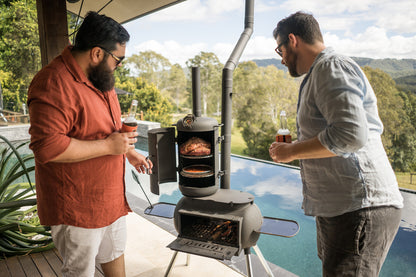 Two men stand by a pool, each holding a drink, grilling meat on an Ozpig Big Pig Smoker Combo barbecue from Ozpig. Trees and hills are visible in the scenic background under a partly cloudy sky.