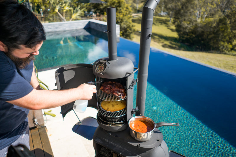 An individual sprays food cooking inside the Ozpig Big Pig Smoker Combo by Ozpig next to a pool. A saucepan with sauce rests on the grill, while trees and grass provide a scenic backdrop—setting the stage for mouthwatering woodfired meals.