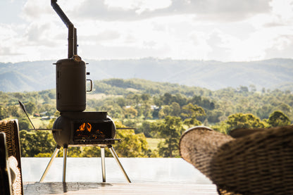 An Ozpig Big Pig Smoker Combo by Ozpig stands on a deck with flames flickering inside its wood-fired chamber and chimney, evoking the promise of delicious woodfired meals. Wicker chairs with a view of green hills and trees under a cloudy sky are arranged to face the smoker.