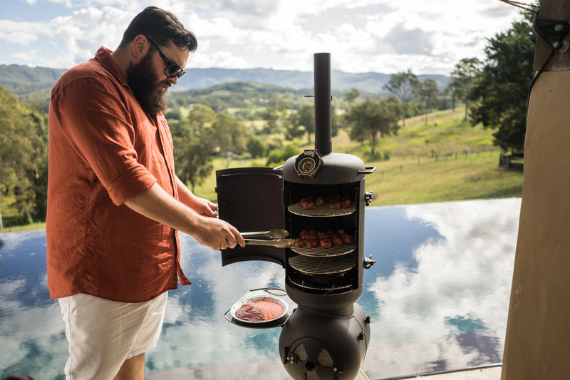A man wearing sunglasses grills meat on an Ozpig Series 2 Smoker Combo by Ozpig beside a pool, with scenic hills and trees in the background under a partly cloudy sky.