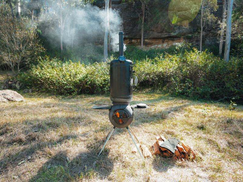 An Ozpig Series 2 Smoker Combo by Ozpig stands on the grass, with smoke rising from its chimney. Firewood is stacked beside this portable outdoor smoker, while trees, bushes, and sunlight filtering through the forest create a scenic backdrop.