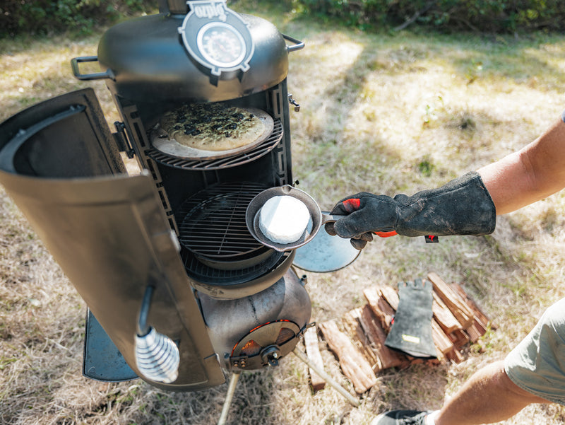 Wearing a heat-resistant glove, someone adds a dish to the Ozpig Series 2 Smoker Combo by Ozpig, where a pizza is already baking on the top rack. Nearby, firewood is stacked on the grass in an outdoor setting, highlighting the versatility of this portable smoker combo.