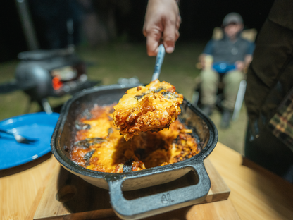 A hand serves a cheesy baked dish, possibly lasagna, from the Campfire Combo Camp Oven outdoors at night, with a blurred person sitting by a fire in the background—perfectly capturing the essence of campfire cooking.