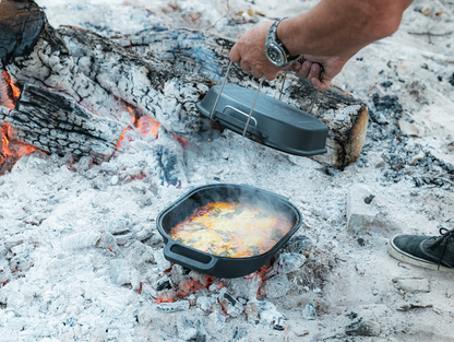 A person lifts the lid off the Campfire Combo Camp Oven, cooking a meal over a campfire surrounded by ash and logs. As steam rises and the food inside is revealed, it captures the essence of traditional campfire cooking.