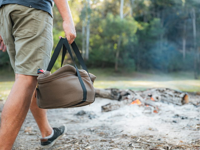 A person in shorts carries the Campfire Combo Camp Oven Canvas Bag, featuring reinforced handles, toward a campfire in a wooded outdoor setting surrounded by trees and grass.