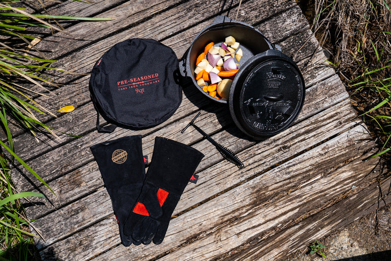 A Campfire Camp Oven Set 9 Quart, pre-seasoned and brimming with chopped vegetables, rests on weathered wood beside its lid, handy carrying case, protective gloves, and a black handle—ideal for outdoor cooking amid grass and lush greenery.