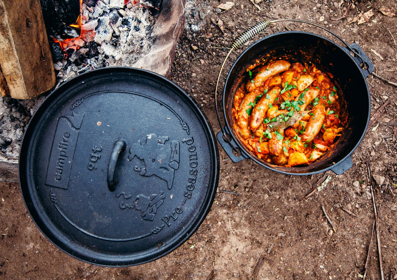A pot of sausage stew with herbs sits on the ground next to its lid, beside a campfire, simmering perfectly in the Campfire 9 Quart Camp Oven—a true delight for outdoor cooking enthusiasts.