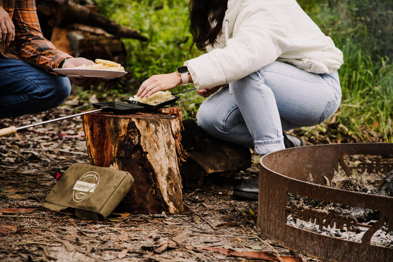 Two people prepare food over a campfire using the Campfire Canvas Bag Jaffle Iron Double Jumbo on the grill. One person crouches to place bread in the jaffle iron, while the other holds a plate. Camping gear—including a sturdy canvas bag—and surrounding greenery complete the scene.