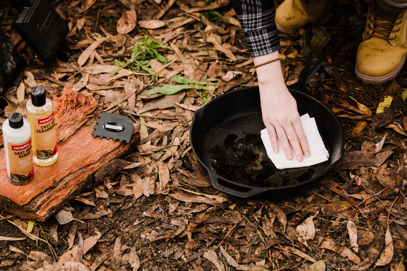 Outdoors on fallen leaves, a person cleans a cast iron skillet with a paper towel. Nearby, Campfire Cast Iron Seasoning Oil, along with a scraper and brush, rests on a wooden board—ready for seasoning the cookware.
