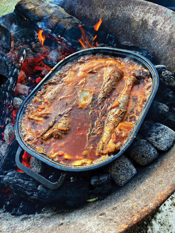 The Campfire Combo Camp Oven, pre-seasoned and ready to use, is filled with fish and vegetables in a red sauce as it cooks over hot coals and burning wood, perfectly showcasing the joys of campfire cooking in an outdoor fire pit.