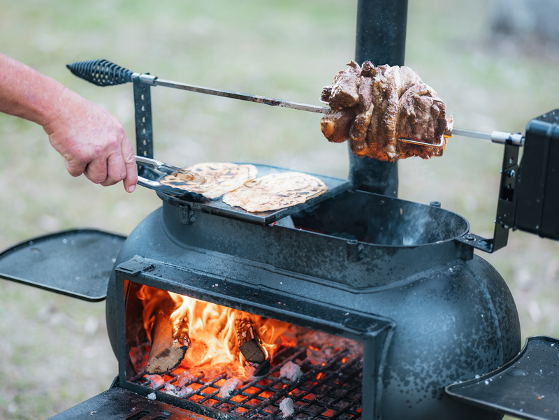A person cooks flatbreads in a pan and roasts meat on a spit over the Ozpig Big Pig outdoor wood-fired stove by Ozpig, with visible flames and logs inside, showcasing the traditional art of woodfired cooking that delivers flavors only an open grill and smoker like the Big Pig can provide.