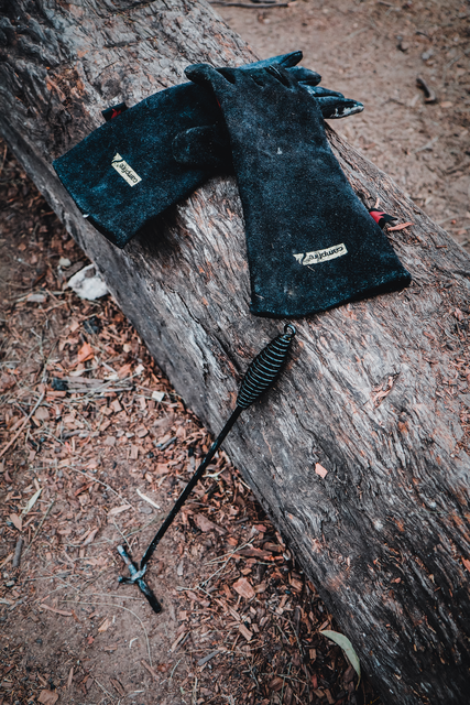 A pair of Campfire Protective Leather Gloves and a metal fire poker rest on a large, weathered log outdoors, surrounded by dirt and scattered leaves—ideal companions for campfire cooking and providing excellent heat insulation during outdoor adventures.