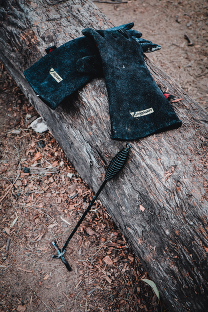 A pair of Campfire Protective Leather Gloves and a metal fire poker rest on a large, weathered log outdoors, surrounded by dirt and scattered leaves—ideal companions for campfire cooking and providing excellent heat insulation during outdoor adventures.