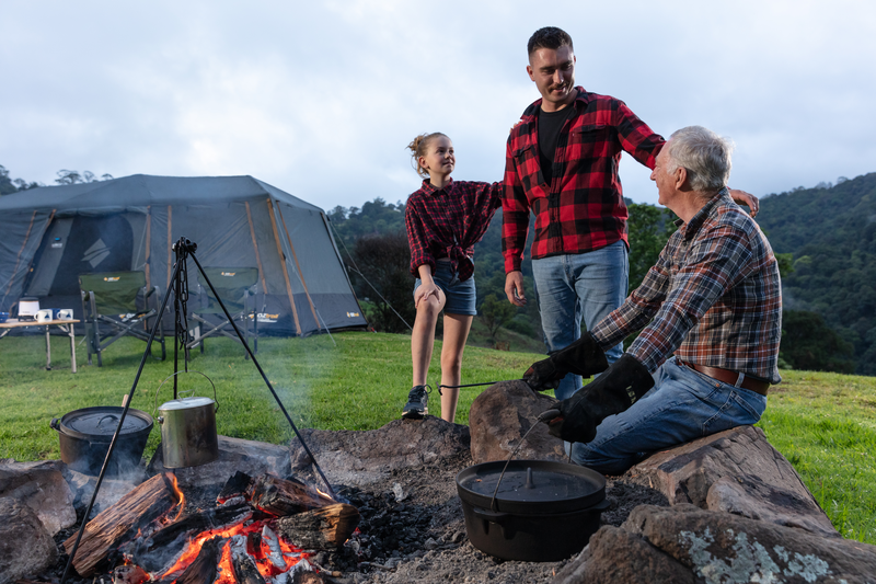 Three people in plaid shirts gather around a campfire at a campsite, with a tent and mountains in the background. A girl, wearing Campfire Protective Leather Gloves for heat insulation, carefully roasts marshmallows while two men, one older, smile and talk. Camping equipment is visible, adding to the cozy scene of campfire cooking under the open sky.