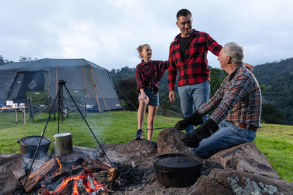 Three people in plaid shirts gather around a campfire at a campsite, with a tent and mountains in the background. A girl, wearing Campfire Protective Leather Gloves for heat insulation, carefully roasts marshmallows while two men, one older, smile and talk. Camping equipment is visible, adding to the cozy scene of campfire cooking under the open sky.