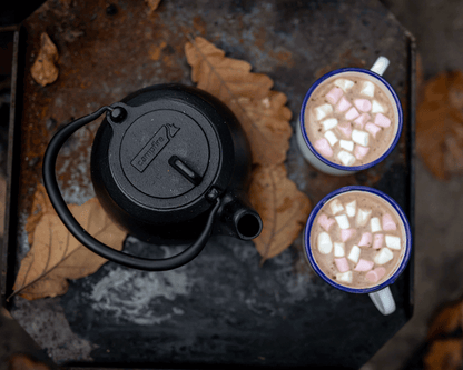 A Campfire 2 Quart Cast Iron Kettle sits on a rustic surface beside two mugs of hot chocolate topped with mini marshmallows, surrounded by scattered brown autumn leaves.