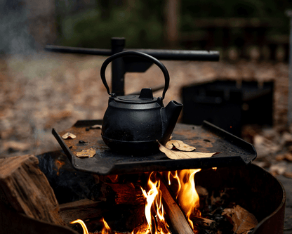 A Campfire 2 Quart Cast Iron Kettle rests on a metal grate above a glowing campfire, flames and logs crackling beneath it. Fallen leaves are scattered on the grate, while an out-of-focus wooded landscape in the background showcases the timeless appeal of this outdoor cookware.