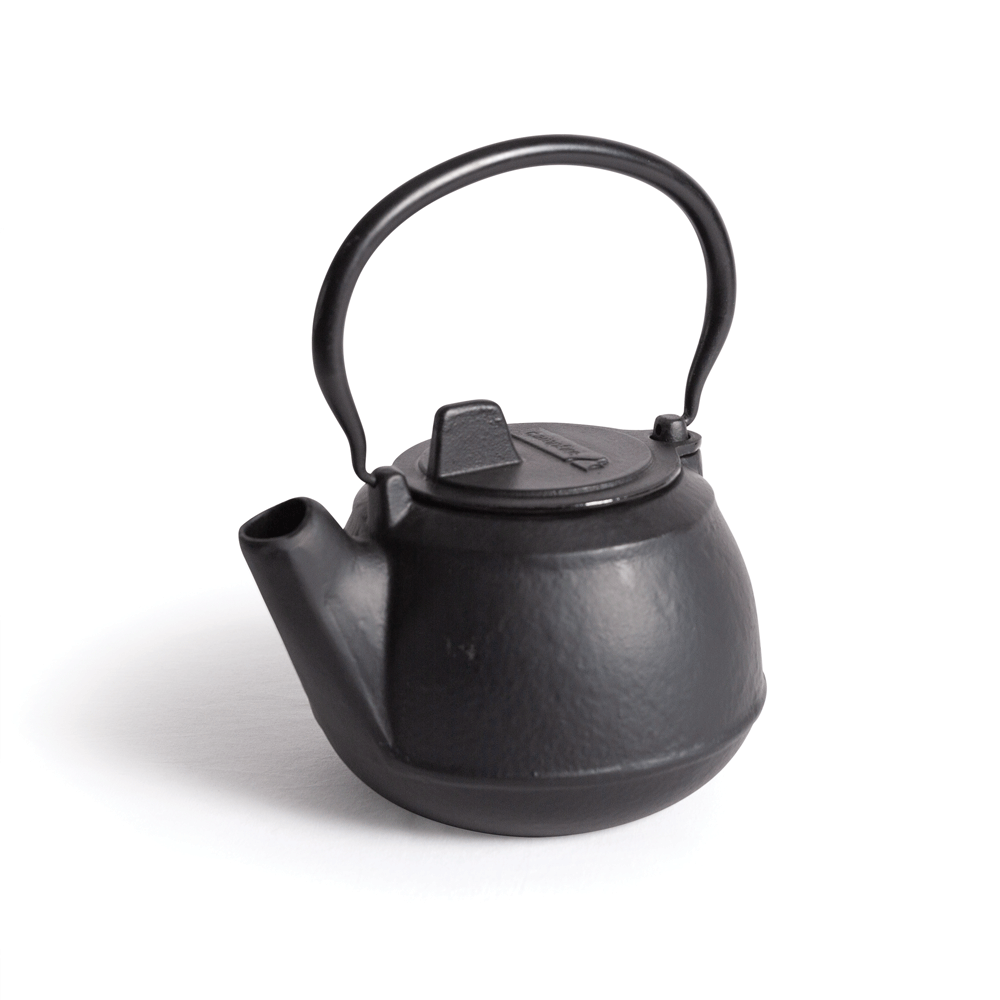 A 2 Quart Cast Iron Kettle by Campfire, featuring a small, black design with a curved handle and short spout, is placed on a white surface.