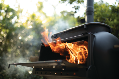 A close-up of the Ozpig Big Pig by Ozpig, an outdoor wood stove with its door open, reveals bright orange flames and smoke within. Surrounded by greenery in the background, this versatile unit functions as both a grill and smoker.