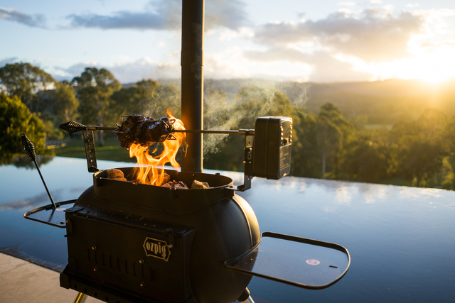 An Ozpig Big Pig by Ozpig is roasting a piece of meat on its rotisserie over an open flame near a pool, with trees and hills visible in the background at sunset—the perfect setup for home woodfired cooking that brings warmth and flavor to outdoor gatherings.