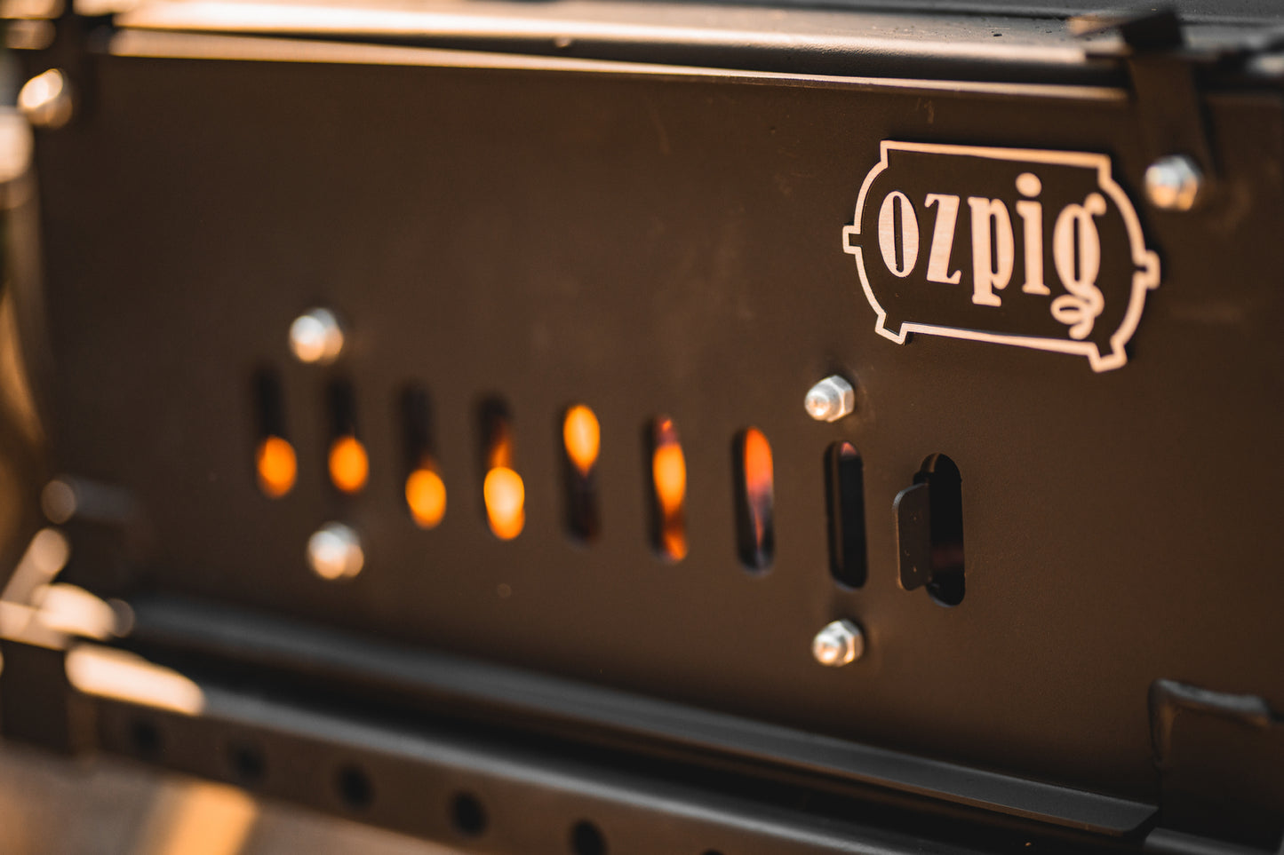 Close-up of the Ozpig Big Pig, a black metal stove from Ozpig, with small vertical openings glowing orange from the fire inside—ideal for outdoor heating or home woodfired cooking. Bolts secure the panel, and the Ozpig logo is visible in the upper right corner.