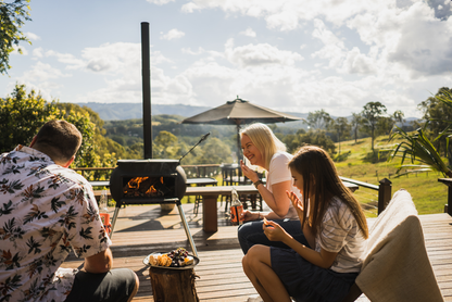 Three people relax on a sunny deck enjoying snacks and drinks beside the Ozpig Big Pig outdoor heating fire pit by Ozpig, with green hills and trees in the background under a partly cloudy sky.