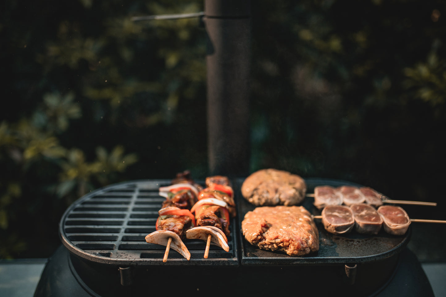 Skewers of vegetables and meat, along with two burger patties and three round meat skewers, cook outdoors on the Ozpig Big Pig Flat Iron BBQ Plate by Ozpig, set against a leafy backdrop that enhances the wood-fired BBQ experience.